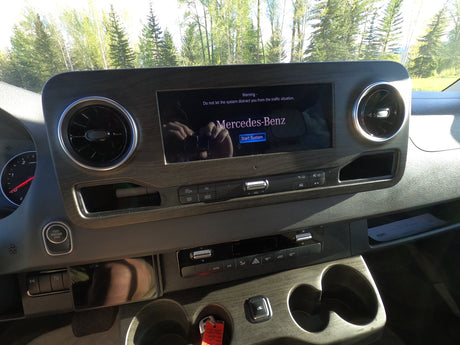 Mercedes-Benz RV rental dashboard: infotainment screen displays a warning. Controls, air vents, and outdoor trees are visible.