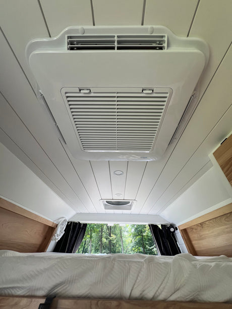 Inside a spacious RV rental, featuring its ceiling-mounted AC unit, vent, and comfortable bed with a forest view.