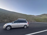 Silver minivan with roof carrier for a road trip, parked by grassy hills. A flexible travel vehicle, perfect for your next RV rental adventure.