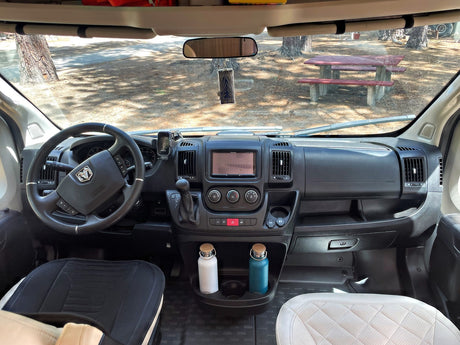 Inside an RV rental: driver's view of the dashboard, steering wheel, and a sunny campsite with a red picnic table.