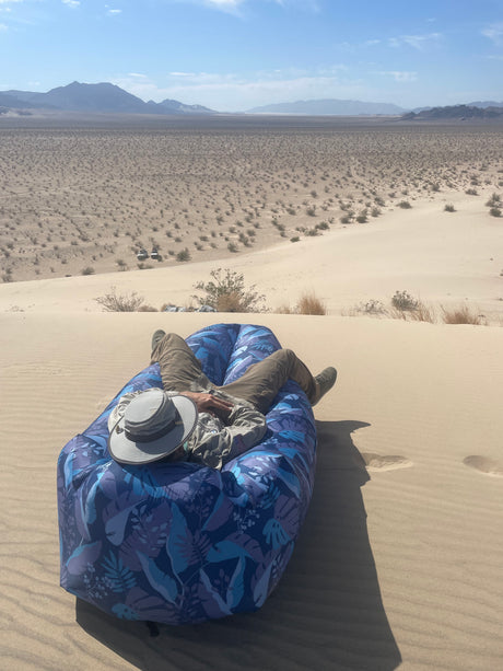 A person lounges on a blue inflatable chair on a sand dune, overlooking a vast desert, mountains, and a distant RV rental.