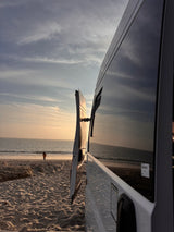 Scenic rv rental parked on a sandy beach. Open van door offers a view of the ocean, soft sunset, and a distant person.
