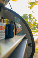 A close-up of coffee supplies inside an rv rental: a white mug, blue mug, metal pot, and a "DRINKING COFFEE" bag on a shelf. Outdoors view.