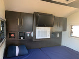 Inside an RV rental bedroom: dual beds, dark wood cabinets, a TV, window AC unit, and a heater for comfort.