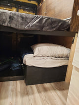 RV rental interior: Wooden walls, black storage, and neatly stored bedding with a blue bag in a compact space.