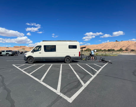 An RV rental camper van parked in a desert lot. A person prepares bikes for outdoor adventure against a scenic backdrop.