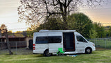 A white Pleasure-Way camper van, a perfect RV rental, parked on a grassy lawn at sunset with a chair and blanket for outdoor leisure.