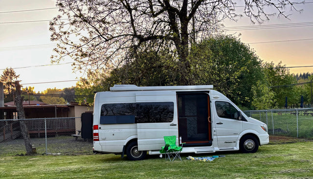 A white Pleasure-Way camper van, a perfect RV rental, parked on a grassy lawn at sunset with a chair and blanket for outdoor leisure.
