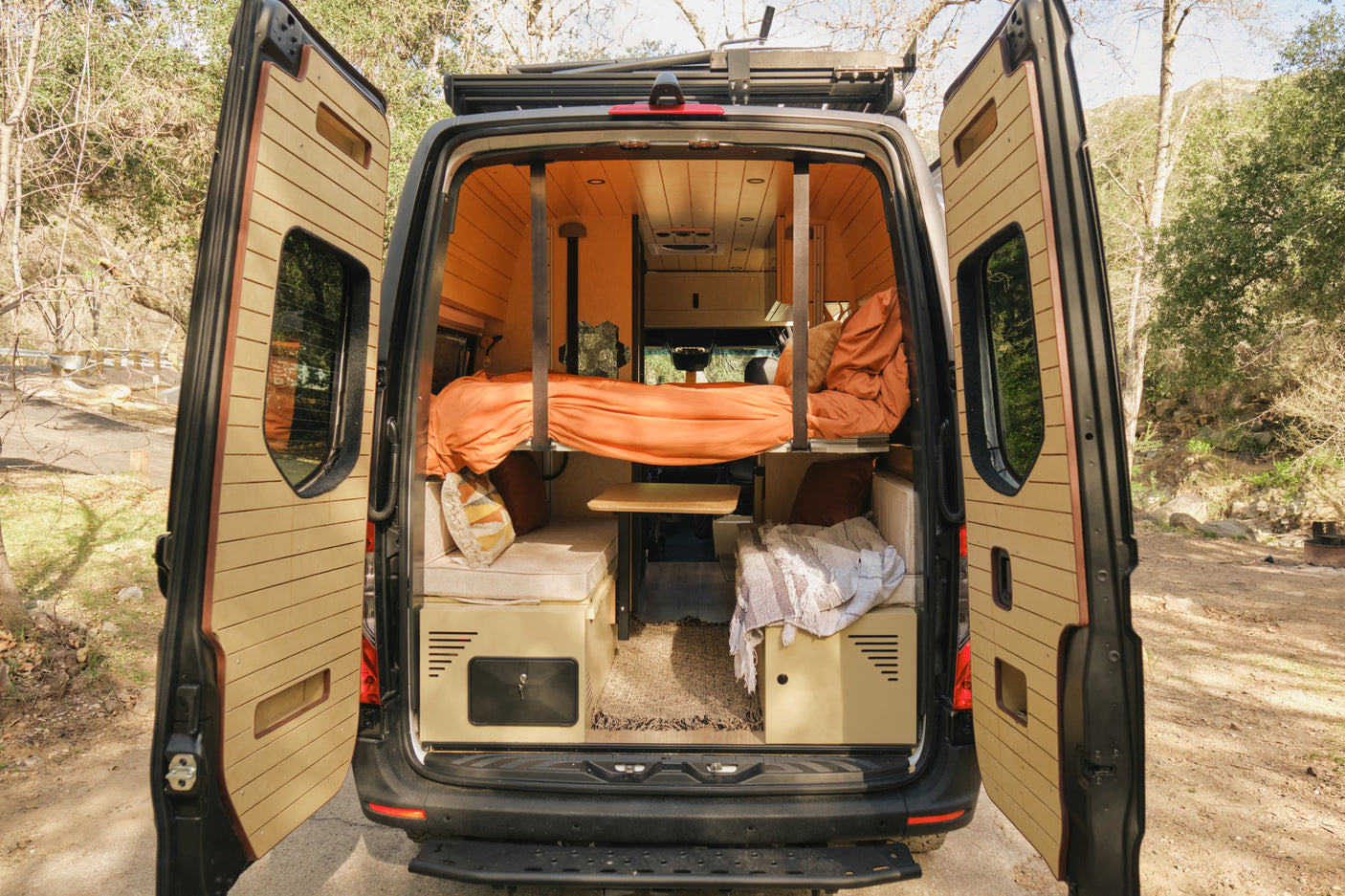 An RV rental's custom wooden interior is seen from open rear doors, featuring a bunk bed with orange bedding, seating, and table.