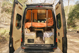 An RV rental's custom wooden interior is seen from open rear doors, featuring a bunk bed with orange bedding, seating, and table.