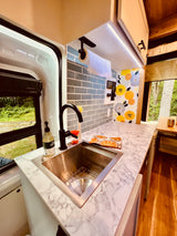 Stylish RV rental kitchen with marble counter, sink, floral backsplash, and a large window view of lush forest.