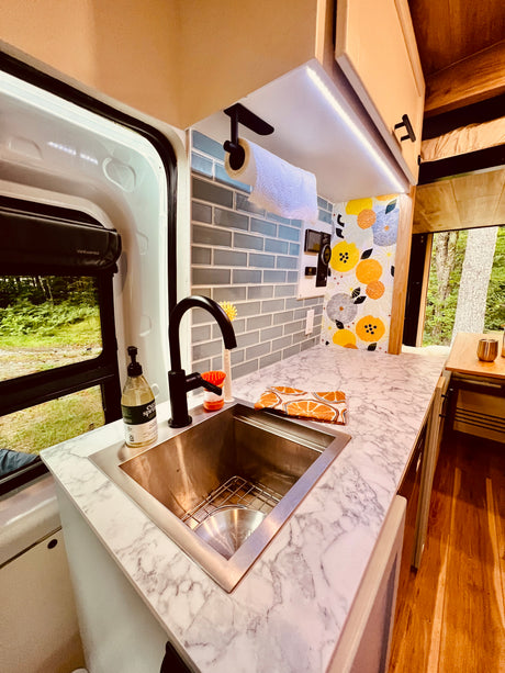Stylish RV rental kitchen with marble counter, sink, floral backsplash, and a large window view of lush forest.