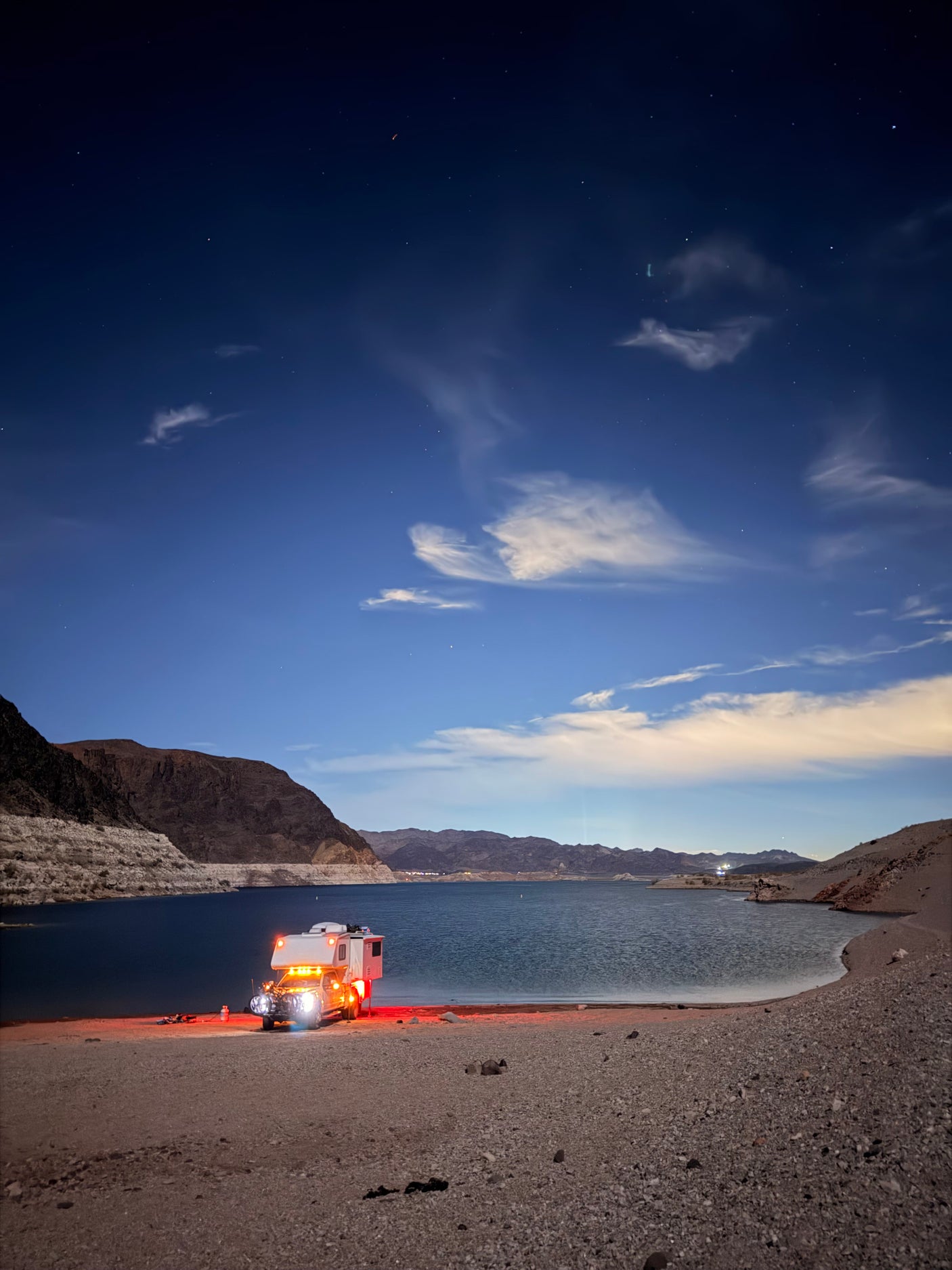 An RV rental lights up a rocky lake beach at night, mountains and a starry sky in the background.