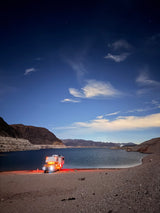 An RV rental lights up a rocky lake beach at night, mountains and a starry sky in the background.
