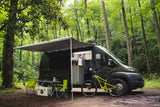 An RV rental camper van parked in a lush forest, featuring an extended awning, outdoor seating, and a mountain bike ready for adventure.
