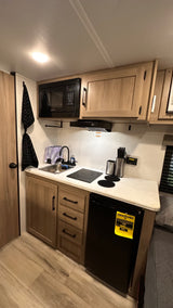 Compact rv rental kitchen with microwave, induction stovetop, sink, and refrigerator. Light wood cabinets and bright lighting.