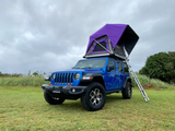 Bright blue Jeep Wrangler with a vibrant purple rooftop tent and ladder, set up on grass. Your RV rental for off-road adventures.