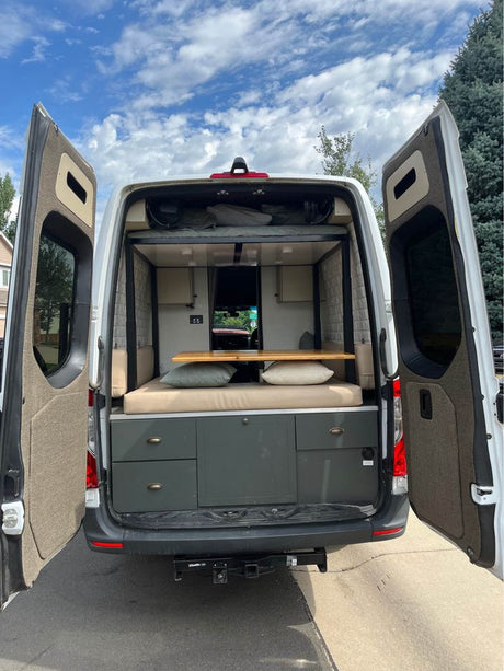 Rear view of an RV rental camper van with open doors, revealing a spacious, custom-built interior featuring bed, table, and storage.