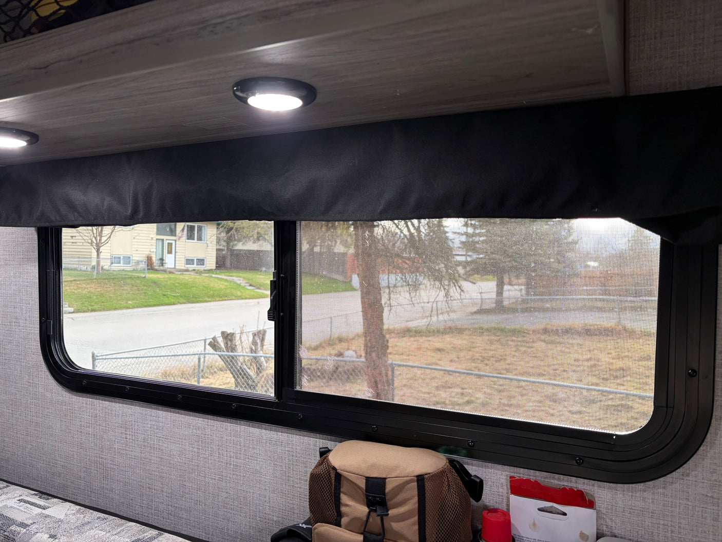 Inside an rv rental: a window with screen reveals a street, house, and trees. Interior features a recessed light, valance, and tan bag.