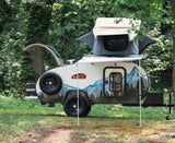 A "Tiny Bug" teardrop RV rental, with a rooftop tent and mountain graphic, sits in a green forest, hatch open.
