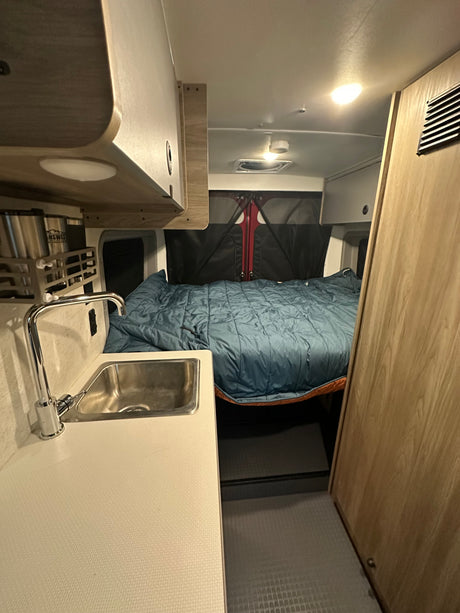 Inside a cozy **rv rental**, showcasing a comfortable bed, compact kitchen with sink, and overhead storage cabinets.