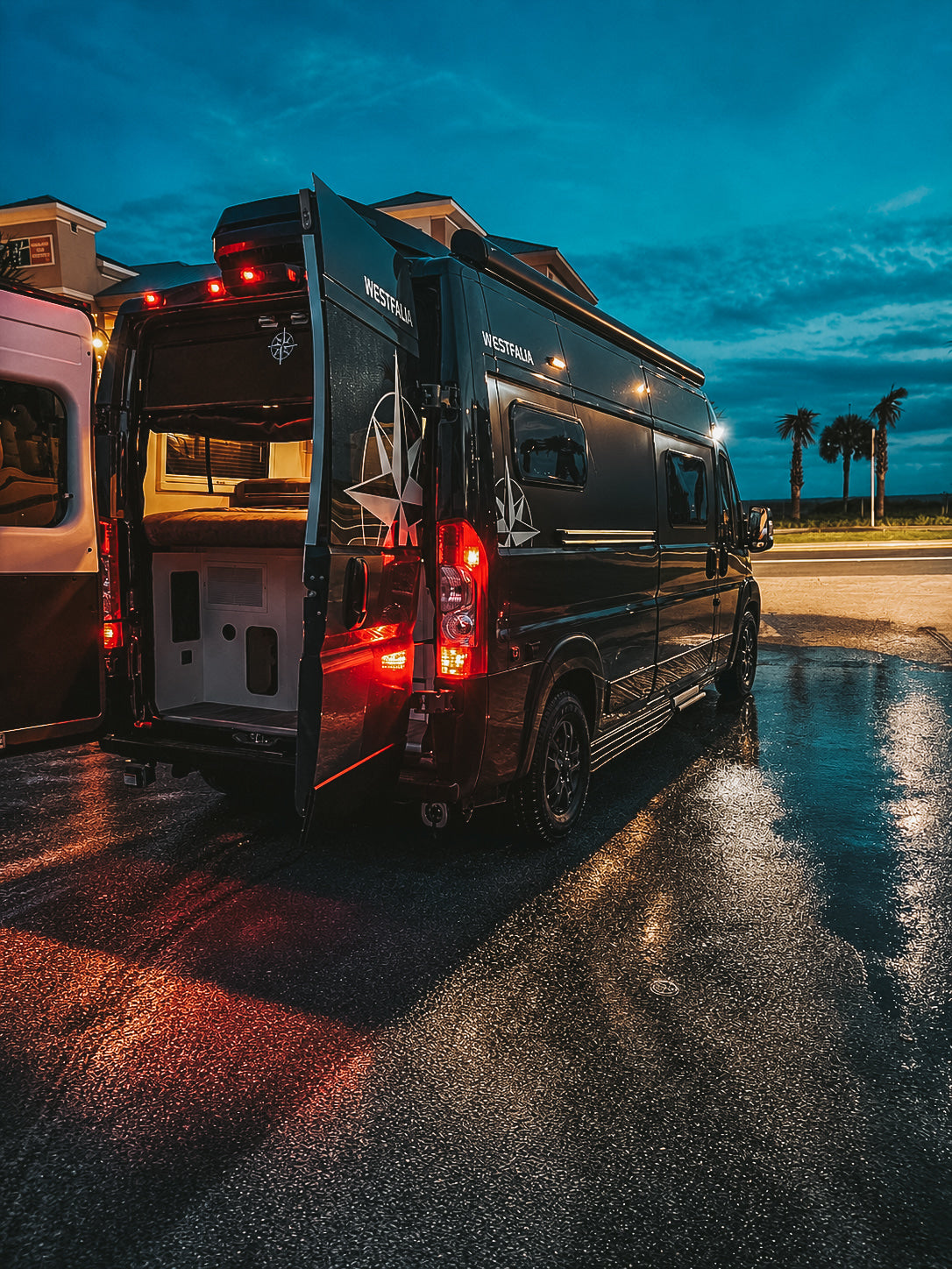 Experience an rv rental! This black Westfalia camper van with open rear doors sits on wet pavement at dusk, near palm trees.