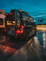 Experience an rv rental! This black Westfalia camper van with open rear doors sits on wet pavement at dusk, near palm trees.