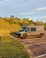Modern grey camper van, perfect for an RV rental adventure, parked on asphalt beside sun-kissed hills and houses.