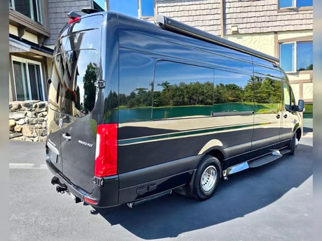 Black luxury Mercedes-Benz Sprinter RV rental. Sleek van parked outside, reflecting a scenic lake & trees.