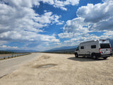 Silver RV rental parked at a scenic mountain overlook. Winding road, distant peaks, and blue sky with white clouds.