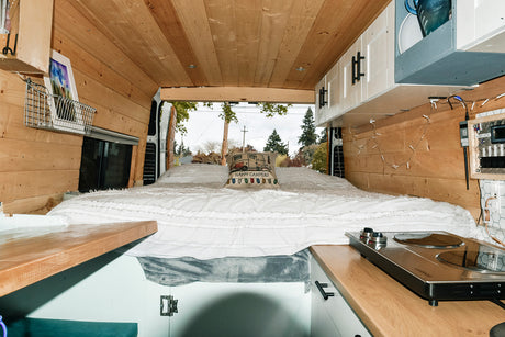 Inside a cozy RV rental: wooden interior, comfy bed with "Happy Campers" pillow, and kitchenette. Back doors open to outdoors.