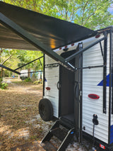 This RV rental travel trailer has its awning extended, with visible entry steps and a spare tire, parked in a sunny campsite.