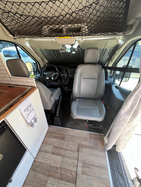 RV rental camper van interior. Front cabin view from living space, showing wood floor, sunshade, and overhead net.