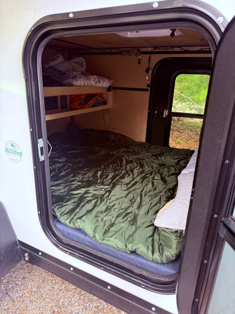 Cozy rv rental interior featuring a large bed, overhead shelves with bedding, and a window offering a view of nature.