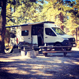 Experience van life with an RV rental! A white Mercedes Sprinter camper van is parked at a shaded forest campsite with an open door and awning.