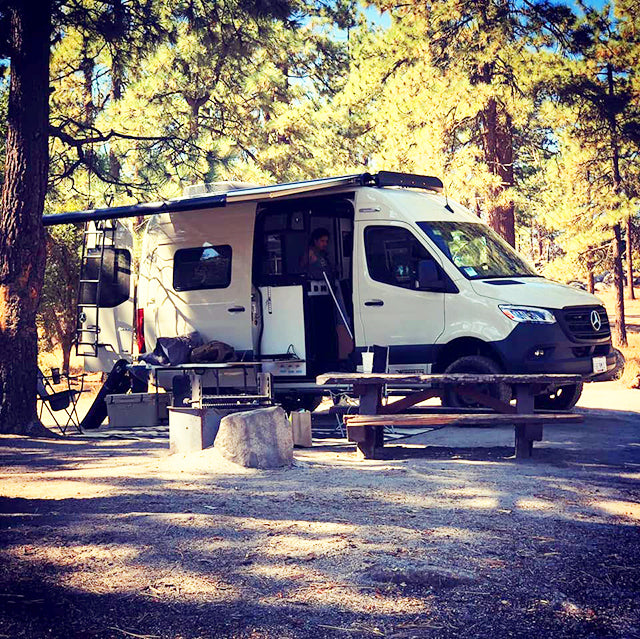 Experience van life with an RV rental! A white Mercedes Sprinter camper van is parked at a shaded forest campsite with an open door and awning.