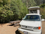 White VW camper van, an rv rental, with pop-top extended, parked by a picnic table in a green, wooded campsite.