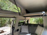 RV rental interior showing a pop-top sleeping area & seating, open to a forest campsite.
