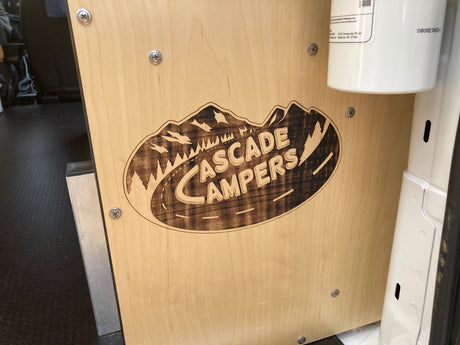 Inside an RV rental: a wooden panel showcases a laser-engraved "Cascade Campers" logo. Screws secure it, with a fire extinguisher nearby.