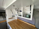 Organized RV rental kitchen & bed area: white overhead cabinets, wood counter, and cozy sleep space.