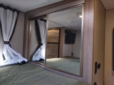 RV rental bedroom interior: green quilted bed, tied-back curtains, and mirrored closet reflecting TV and fridge setup.