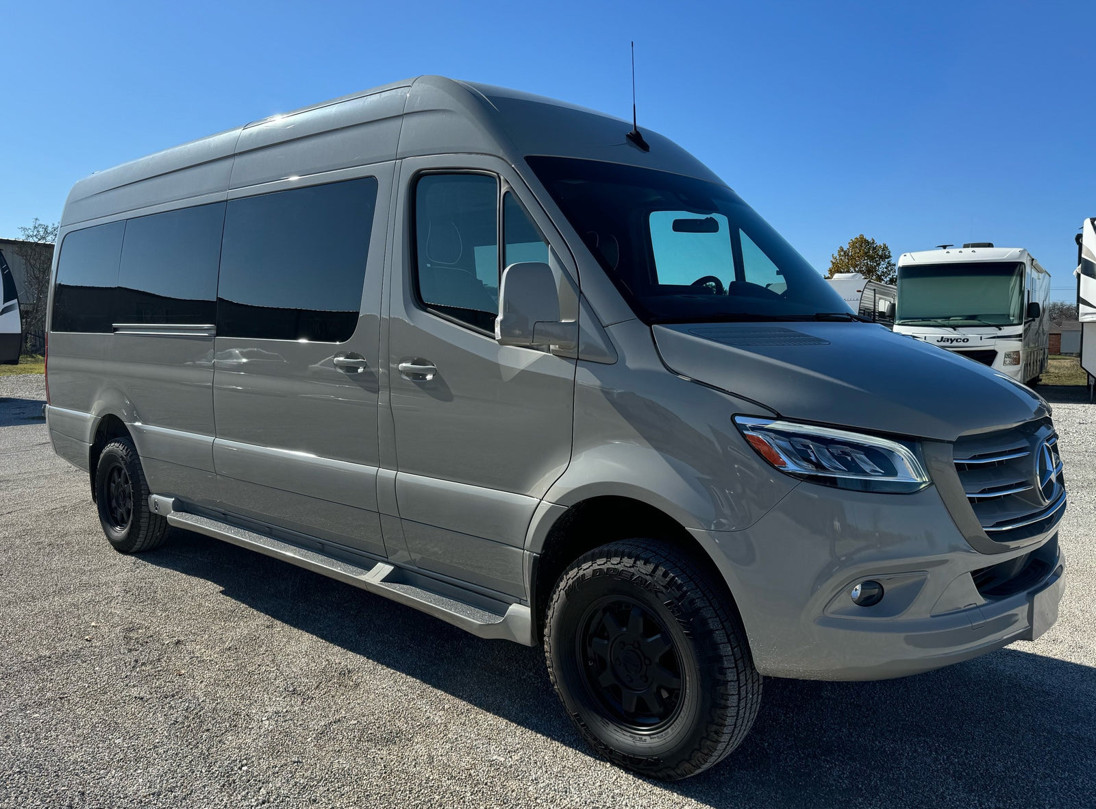 A sleek grey Mercedes-Benz Sprinter van, ready for rv rental, parked outdoors on gravel. Other RVs are in the background.