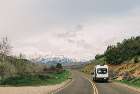 White rv rental van on a scenic road, flanked by green hills, with snow-capped mountains in the distance under a cloudy sky.
