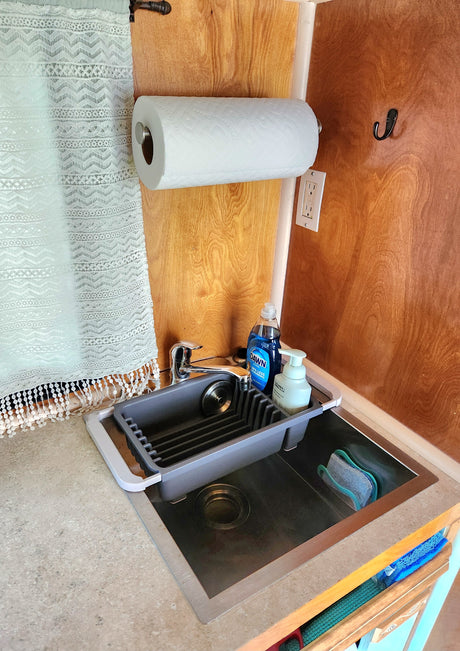 An RV rental kitchen sink area: stainless steel sink with faucet, dish soap, hand soap, and drying rack. Paper towels and outlet on wood wall.