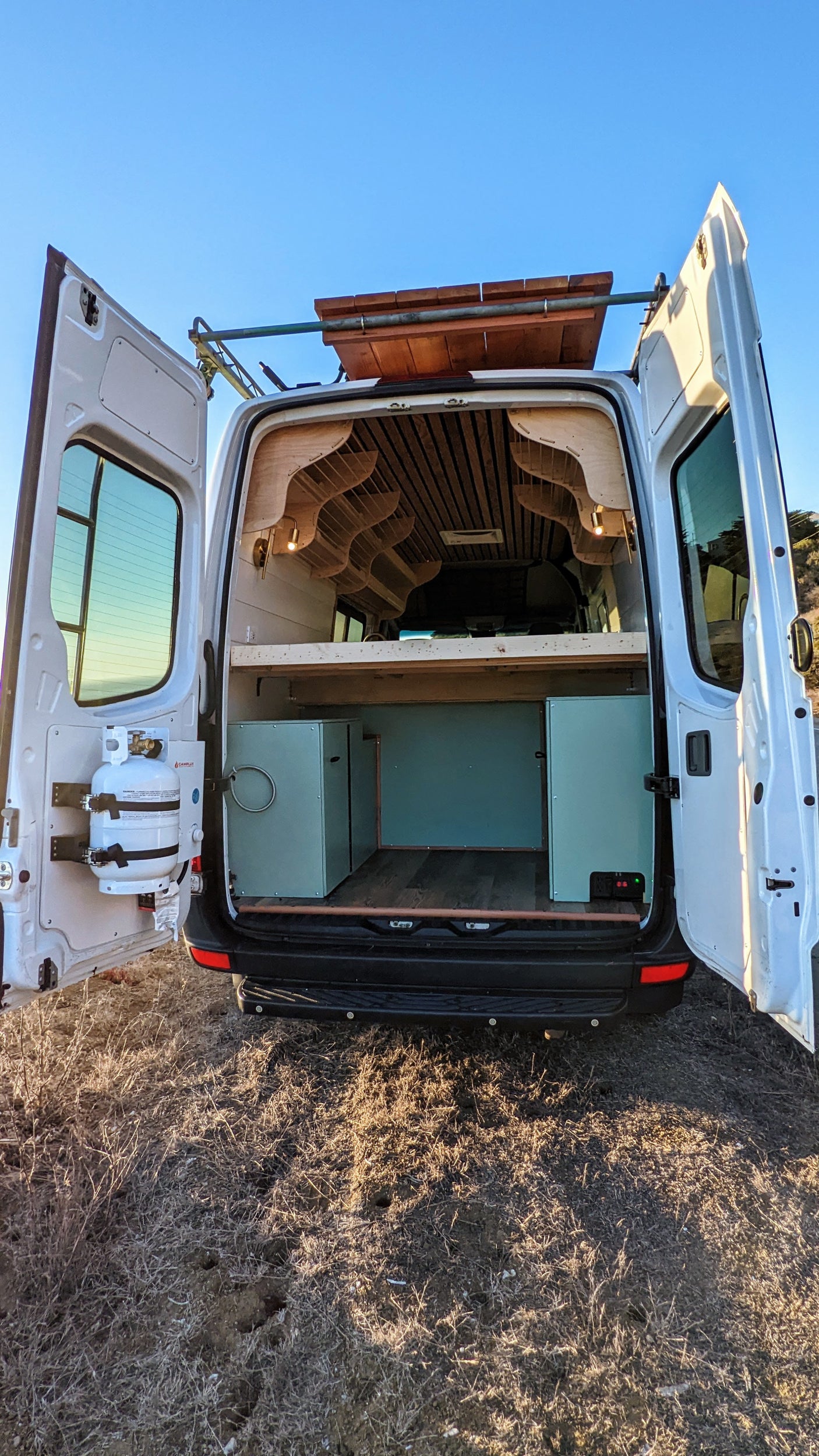 An RV rental camper van with open rear doors reveals a custom wood interior, bed, and storage. Ideal for road trips.
