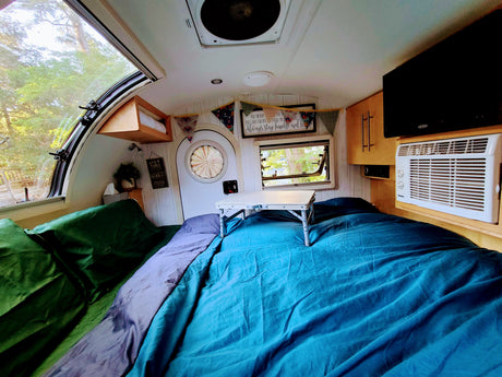 Cozy RV rental interior with a large bed, a bed table, AC unit, and windows showcasing a serene forest view.