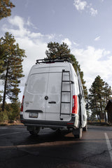 This white Mercedes Sprinter van, perfect for an RV rental adventure, features a roof rack & ladder, parked in a scenic pine forest.