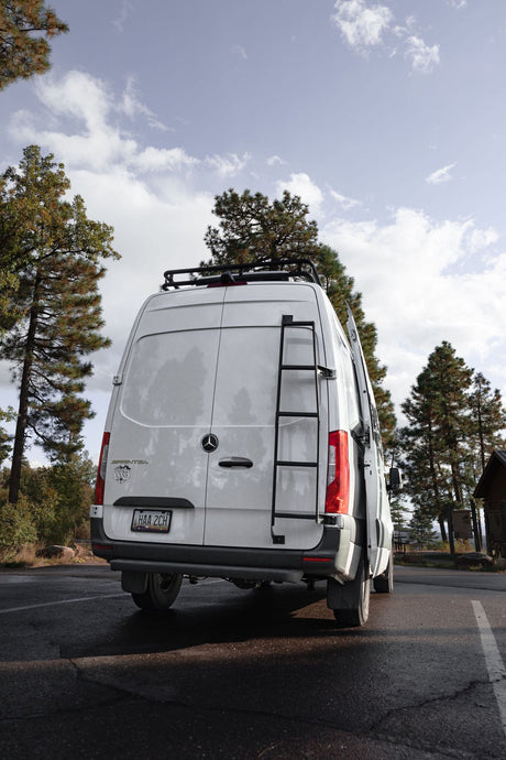 This white Mercedes Sprinter van, perfect for an RV rental adventure, features a roof rack & ladder, parked in a scenic pine forest.