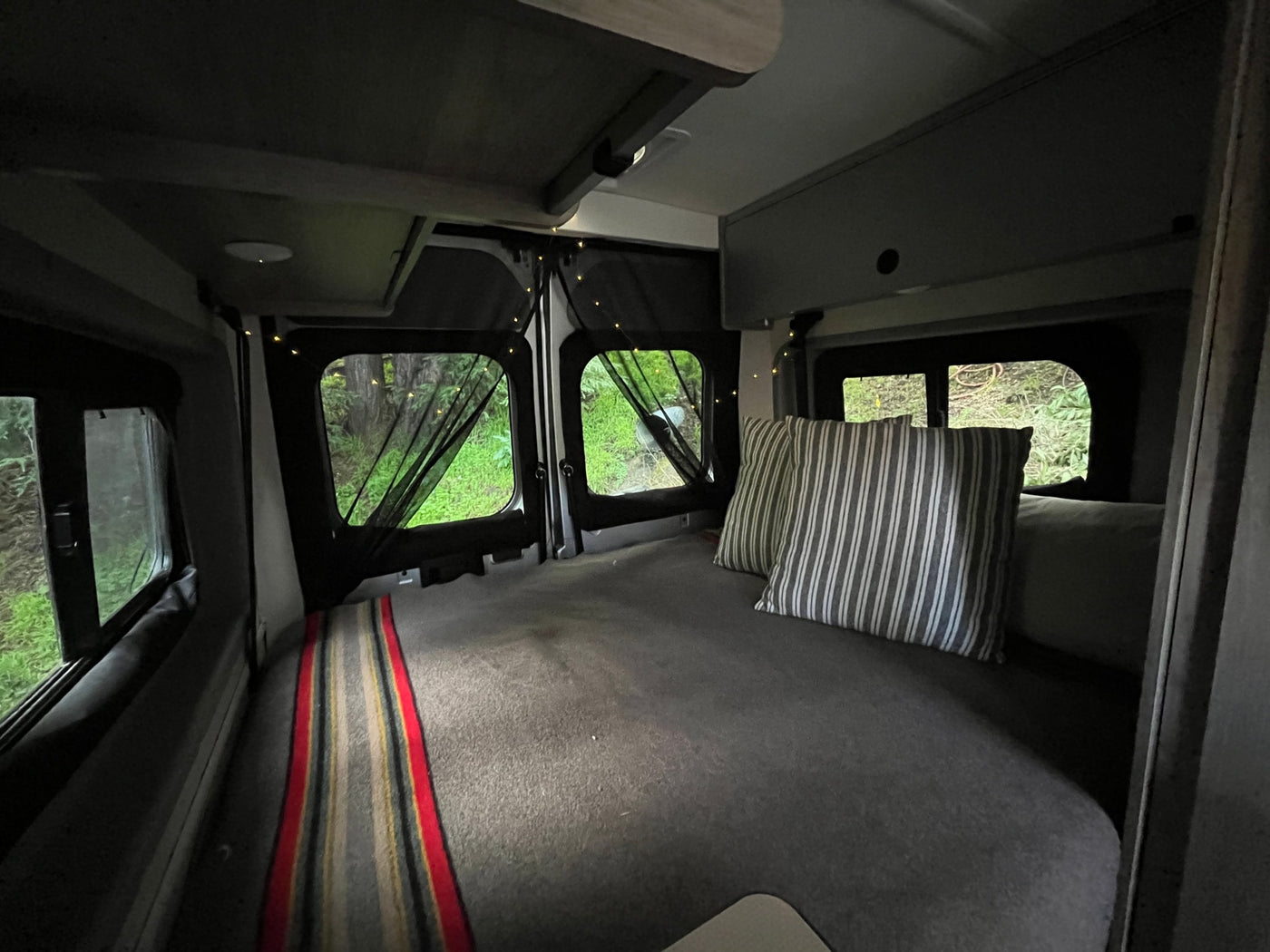 Relax in this cozy RV rental's camper van bedroom. Features a comfortable bed with striped pillows & natural views with string lights. — photo 20