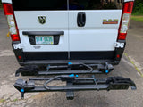 RV rental: Rear of a white Ram ProMaster van with a two-tier bicycle hitch rack, ready for adventure on a wet day.
